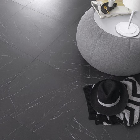 Un sol en carrelage gris foncé à motifs de marbre supporte un pouf gris avec des livres et une lampe. Un chapeau noir et un tiss