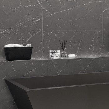 A black rectangular bathtub sits against gray marble walls. above, a shelf holds a basket with towels, a decorative box, and a r