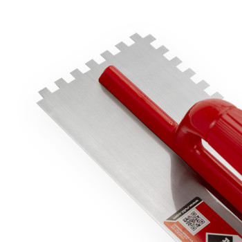 Uma espátula dentada com cabo vermelho repousa sobre uma superfície branca. Possui lâmina de metal com entalhes quadrados e incl