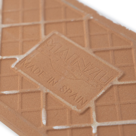 Um ladrilho retangular de cor terracota apresenta texto em relevo que diz `MANUAL` e `MADE IN SPAIN`, com um padrão de grade e a