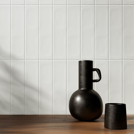 Um vaso e uma xícara pretos foscos repousam sobre uma superfície de madeira, encostados em uma parede de azulejos brancos, lança