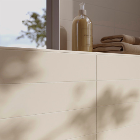 Azulejos de cor creme revestem a parede do banheiro, lançando sombras suaves. Perto dali, um dispensador de sabonete com a etiqu