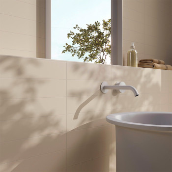 An einer glatten, beige gefliesten Wand ragt eine Badewannenarmatur hervor. Sonnenlicht strömt durch ein Fenster und wirft Baums