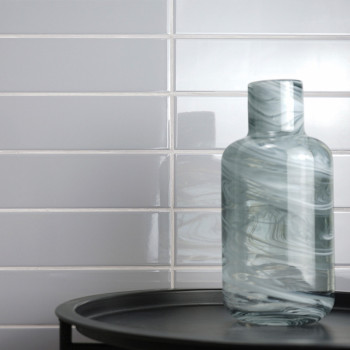 Een gewervelde glazen fles rust op een zwart dienblad, tegen een glanzende witte metrotegelmuur, en reflecteert het omgevingslic