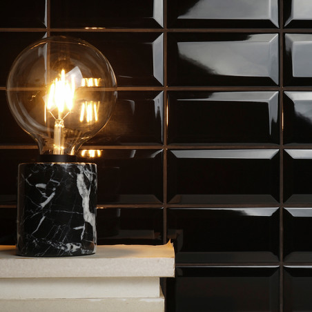 Una bombilla incandescente se asienta sobre una base de mármol negro, iluminando un fondo de pared de azulejos negros brillantes Una bombilla incandescente se asienta sobre una base de mármol negro, iluminando un fondo de pared de azulejos negros brillantes