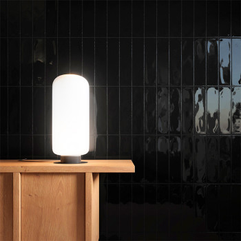 Une lampe haute et cylindrique émet une lumière douce sur une table en bois, posée contre un mur carrelé noir brillant réfléchis