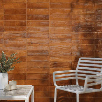 Un mur de carreaux rectangulaires marron brillants reflète la lumière, situé derrière une chaise et une table en métal blanc, or