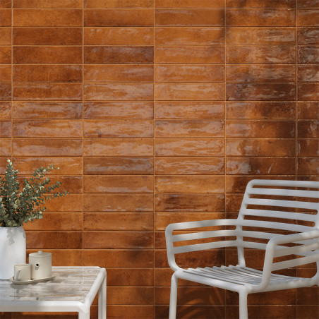 Una pared de azulejos rectangulares de color marrón brillante refleja la luz, situada detrás de una silla y una mesa de metal bl