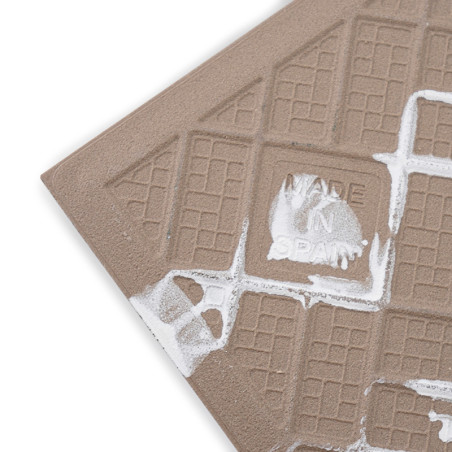 Un azulejo de cerámica marrón texturizado está grabado con patrones de cuadrícula y salpicaduras de pintura blanca, y presenta e