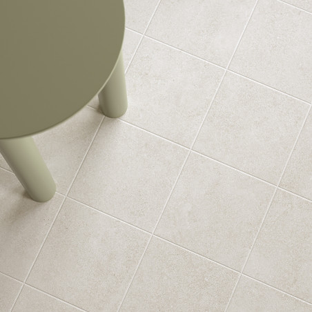 Uma perna de mesa redonda verde-oliva fica sobre uma grade de azulejos bege claro, criando uma aparência de piso moderna e minim