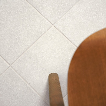 Uma grade de ladrilhos cinza claro é plana, com uma perna de cadeira de madeira entrando na moldura pela direita, sugerindo piso