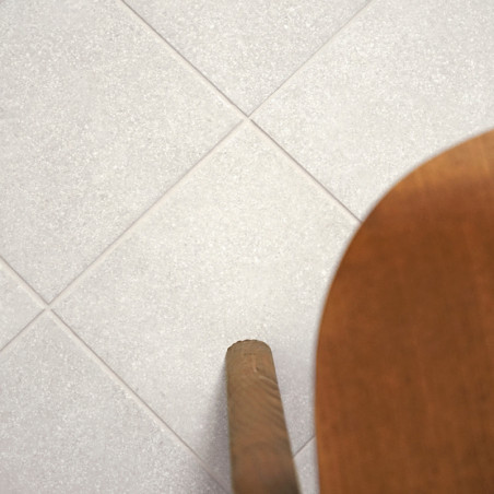 Uma grade de ladrilhos cinza claro é plana, com uma perna de cadeira de madeira entrando na moldura pela direita, sugerindo piso