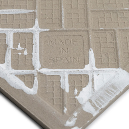 A beige tile with embossed grid patterns features `MADE IN SPAIN` text, partly covered by white grout, against a neutral backdro