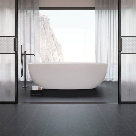 A white freestanding bathtub stands centered in a minimalist bathroom with black tiles, framed by large windows revealing a clif