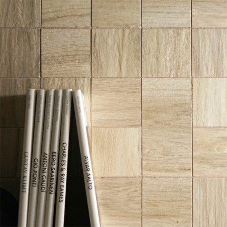 Helle Holzfliesen bilden ein Gittermuster an einer Wand, nach links lehnen sich mehrere vertikal stehende Bücher. Zu den Titeln 