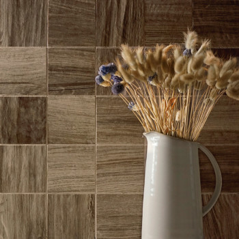 Een witte keramische kan bevat gedroogde beige en paarse bloemen, tegen een muur van bruine tegels met houtstructuur.