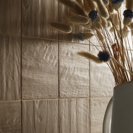 Des carreaux beiges texturés recouvrent un mur, reflétant une lumière douce. Un vase blanc contient des fleurs séchées, brun ten