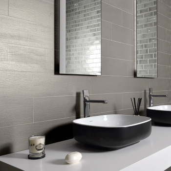 A modern bathroom sink is set within a sleek countertop, accompanied by a soap bar and candle, against a gray tiled wall with mi