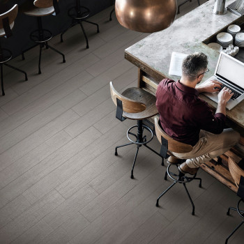 Uma pessoa, sentada em uma banqueta, digita em um laptop no balcão da cozinha. A sala dispõe de bancos de madeira e piso de made