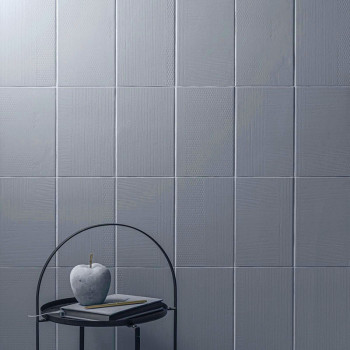 Graue quadratische Fliesen bedecken eine Wand und erzeugen eine strukturierte Oberfläche. Vorne auf einem Metalltisch stehen ein