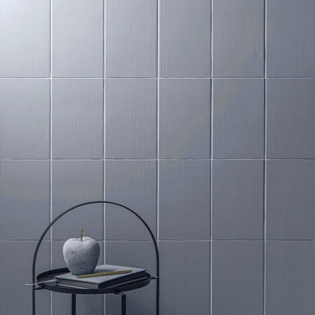 Graue quadratische Fliesen bedecken eine Wand und erzeugen eine strukturierte Oberfläche. Vorne auf einem Metalltisch stehen ein