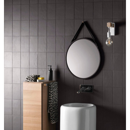 Un miroir rond est suspendu au-dessus d’un lavabo cylindrique blanc dans une salle de bains aux murs carrelés sombres. Une armoi