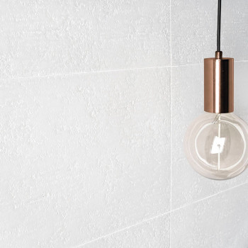 Uma parede de azulejos brancos texturizados é acompanhada por uma lâmpada suspensa com soquete de cobre, pendurada em um ambient