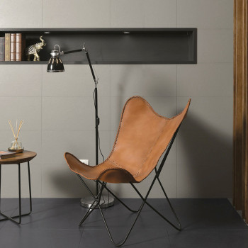 A leather butterfly chair sits on dark tile flooring, next to a modern floor lamp and small round table. A shelf with decor is s