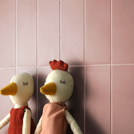 Dos figuras de patos de peluche están apoyadas contra una pared de azulejos rosas, ambos con los ojos cerrados y vestidos con tr