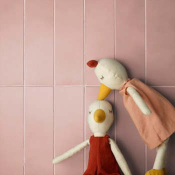 Twee speelgoedvogels met gesloten ogen en oranje snavels leunen tegen een zachtroze betegelde muur en dragen rode en roze outfit