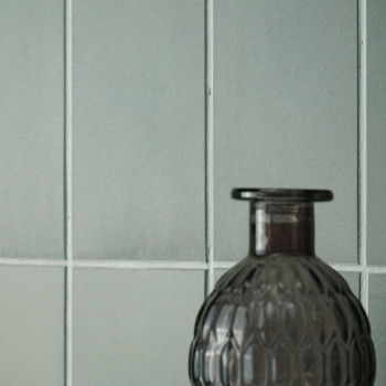 Une bouteille en verre gris texturé se dresse sur un fond de carreaux lisses vert pâle disposés selon un motif en grille.
