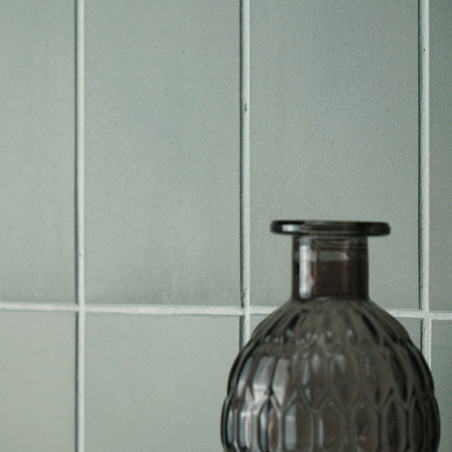 Una botella de vidrio gris texturizada se alza sobre un fondo de azulejos lisos de color verde pálido dispuestos en forma de cua