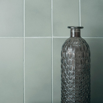 Una botella alta de vidrio texturizado se alza sobre un fondo de azulejos cuadrados de color gris claro mate, lo que sugiere una