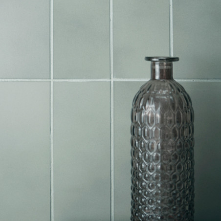 Une grande bouteille en verre texturé se dresse sur un fond de carreaux carrés gris clair mat, suggérant une esthétique minimali