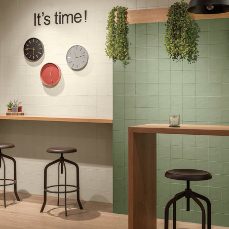 Hocker stehen an Holztheken in einem gefliesten Raum mit Hängepflanzen. An der Wand hängen drei Uhren mit der Aufschrift: „Es is