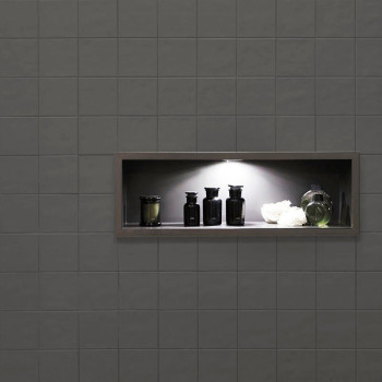 Uma prateleira embutida com garrafas pretas e um vaso verde sob iluminação suave fica dentro de uma parede de azulejos cinza esc
