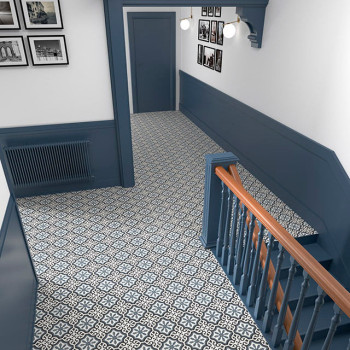 Gemusterte geometrische Fliesen bedecken einen Flurboden, der von einer blauen Täfelung und einer Treppe begrenzt wird. Monochro