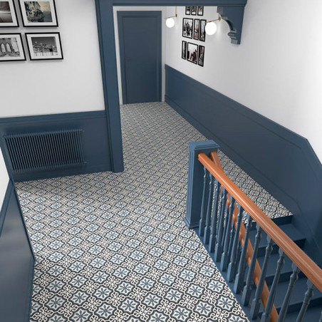 Gemusterte geometrische Fliesen bedecken einen Flurboden, der von einer blauen Täfelung und einer Treppe begrenzt wird. Monochro