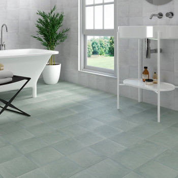A bathroom features matte gray tiles on the floor, a freestanding white bathtub, a modern sink, and a tall potted plant by a win