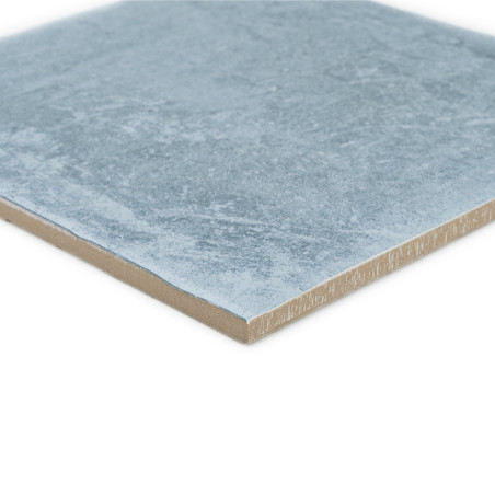 Vierkante keramische tegel met een lichtblauw gestructureerd oppervlak, plat liggend. De rand onthult een lichtbruine laag.