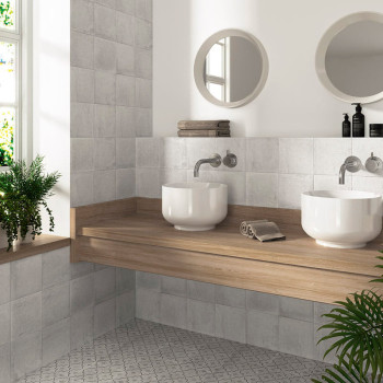 Deux lavabos blancs reposent sur un comptoir en bois, entourés de murs et de sols carrelés gris, avec des miroirs ronds et de la