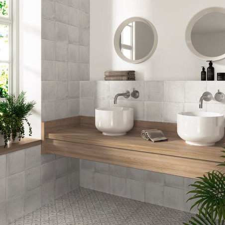 Dos lavabos blancos descansan sobre una encimera de madera, rodeados de paredes y piso de azulejos grises, con espejos redondos 