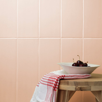 Une table en bois contient un bol de cerises blanches, orné d'un tissu à carreaux rouges, sur fond de murs carrelés roses.