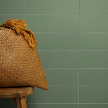 Una cesta tejida con borlas se encuentra sobre un taburete de madera sobre un fondo de azulejos verdes y lisos.