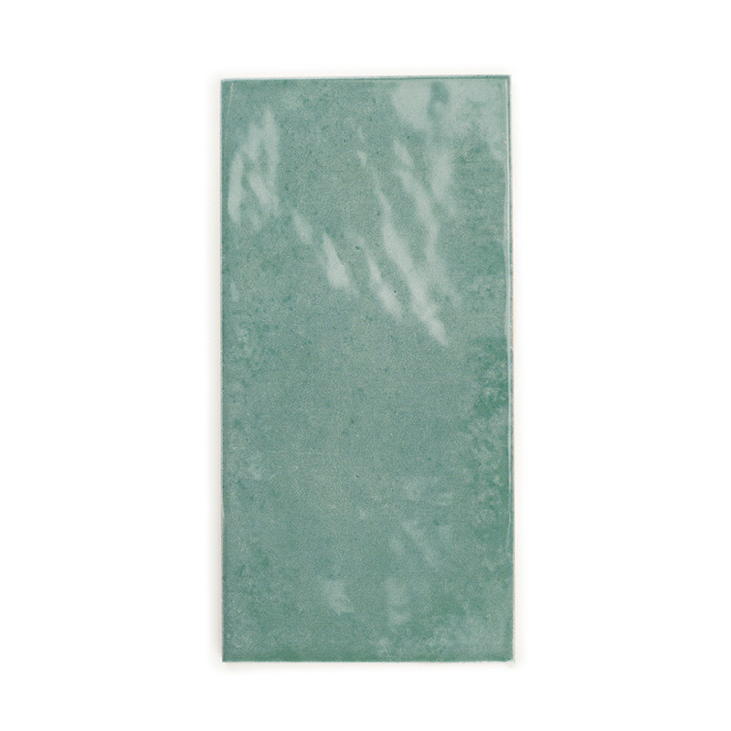 Azulejo cerámico rectangular con una superficie verde brillante que refleja ligeramente la luz. Se apoya sobre un fondo blanco l
