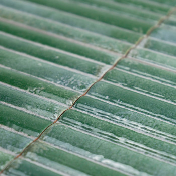 Ladrilhos verdes com acabamento brilhante, dispostos em fileiras paralelas, refletem a luz. Suas texturas de superfície criam va