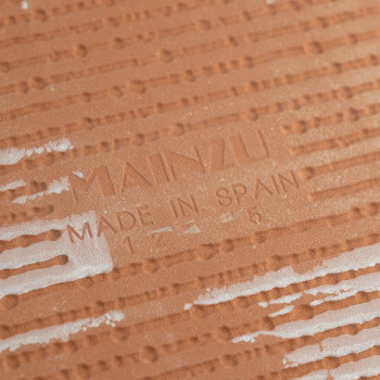 Um ladrilho de terracota, texturizado com linhas horizontais em relevo e manchas brancas, exibe o texto `MAINZU MADE IN SPAIN` n