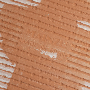 Un carreau de céramique brun orangé, texturé de motifs linéaires. Le texte « MAINZU MADE IN SPAIN » est estampé sur la surface.