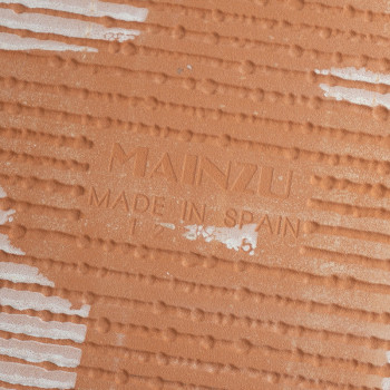 The terracotta tile is embossed with `Mainzu Made in Spain,` featuring diagonal ridged patterns and interspersed white streaks t