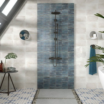 A central dark showerhead mounted on vertical blue-striped tiles sits amidst beige wall tiles. Around, there are towels, a plant